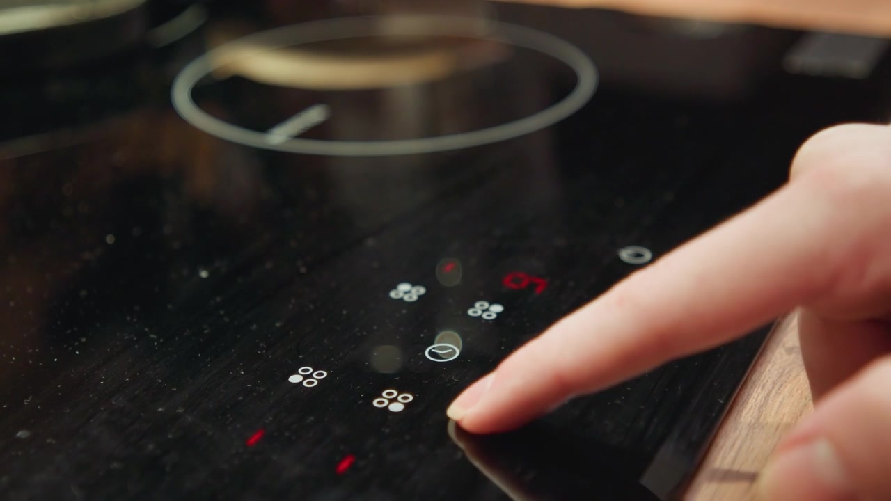 Una vista ravvicinata mostra un dito indice che preme un controllo touch luminoso su un piano cottura a induzione nero lucido, con una zona di riscaldamento circolare, una superficie in vetro riflettente e un piano di lavoro in legno che creano un ambiente pulito e moderno.
