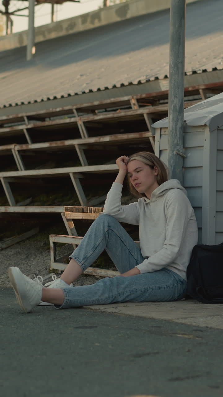 молодая женщина сидит вдумчиво на тротуаре стадиона, опираясь головой на руку с черным пакетом рядом с ней, пустые трибуны создают спокойный, отражающий фон