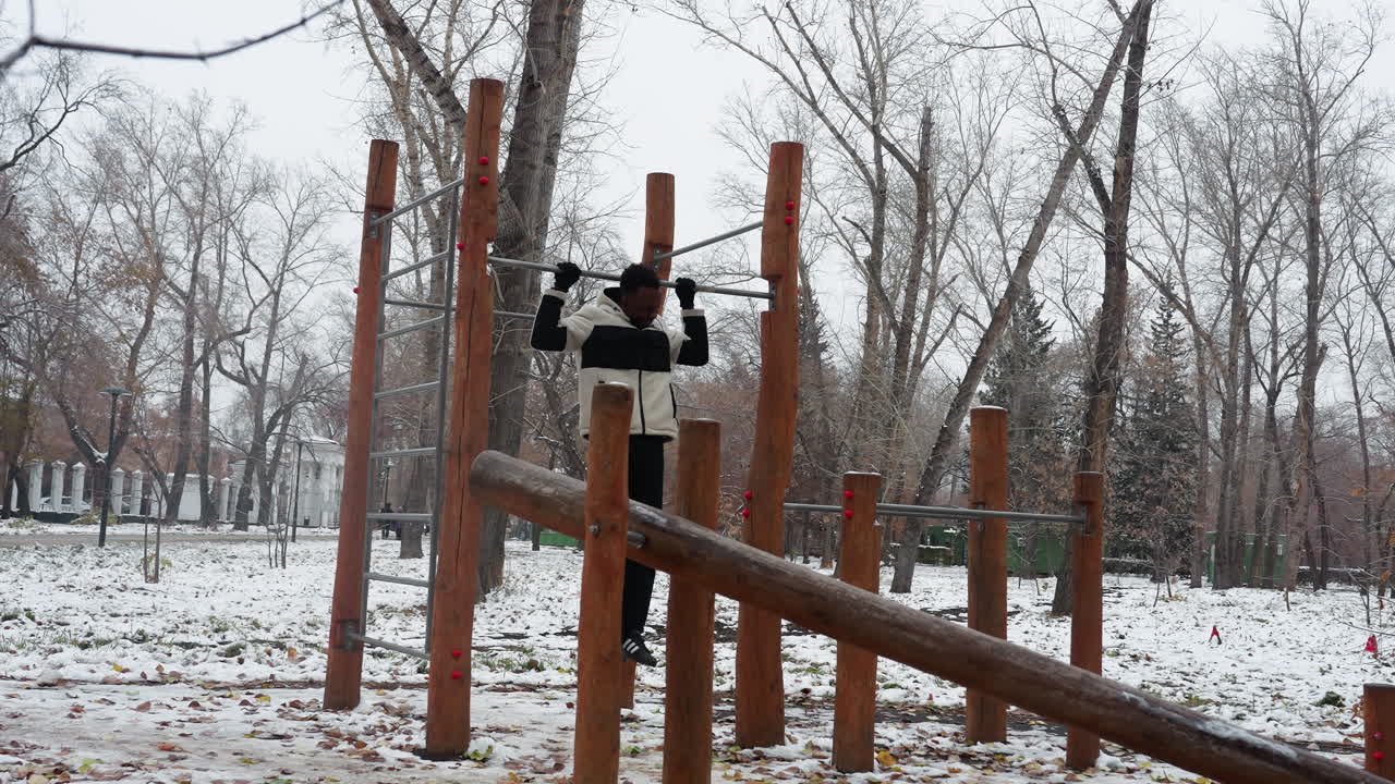 молодой взрослый активно занимается отжиманиями на открытом тренажерном оборудовании зимой, окруженный голыми деревьями в заснеженном парке, иллюстрируя приверженность фитнесу, несмотря на холод