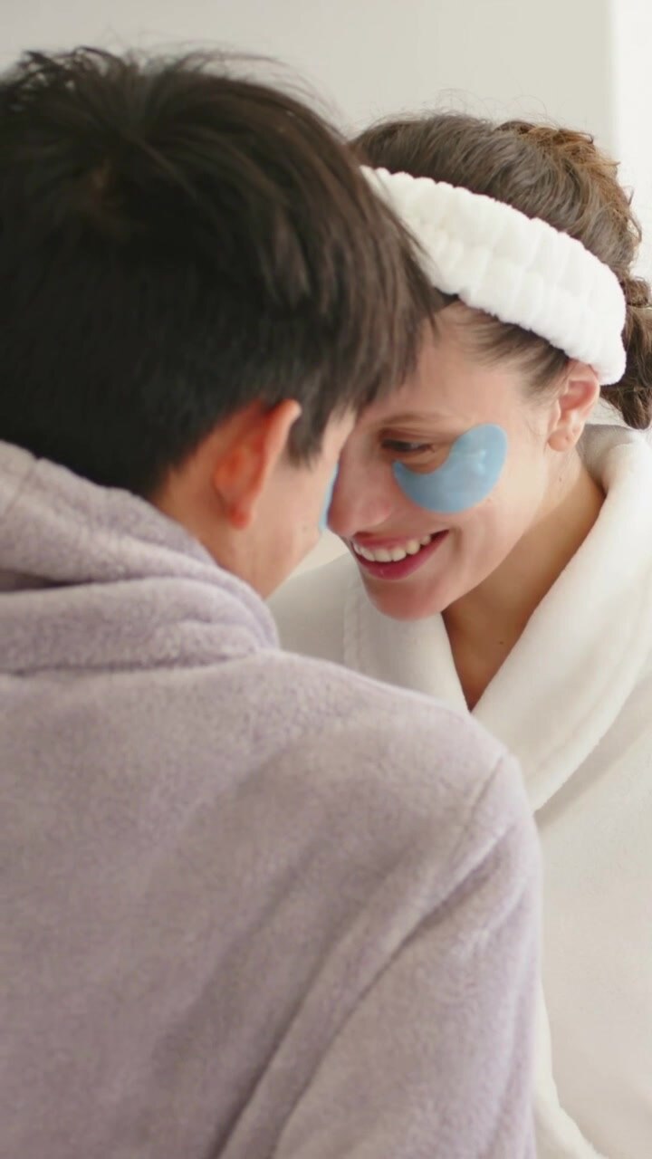 Vertical video: Smiling couple leaning closer at home for skincare, wearing eye patches and robes
