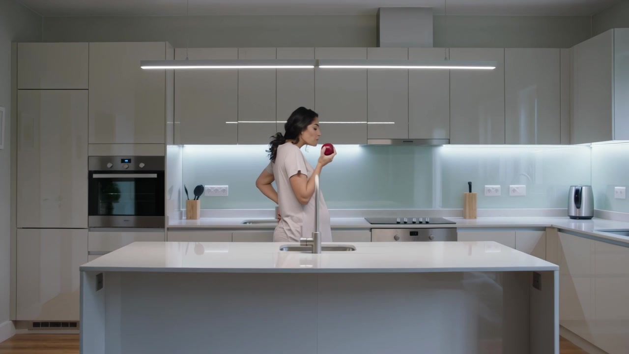 Mulher grávida com vestido solto e leggings, segurando uma maçã vermelha nos lábios, lanchando na cozinha de casa.