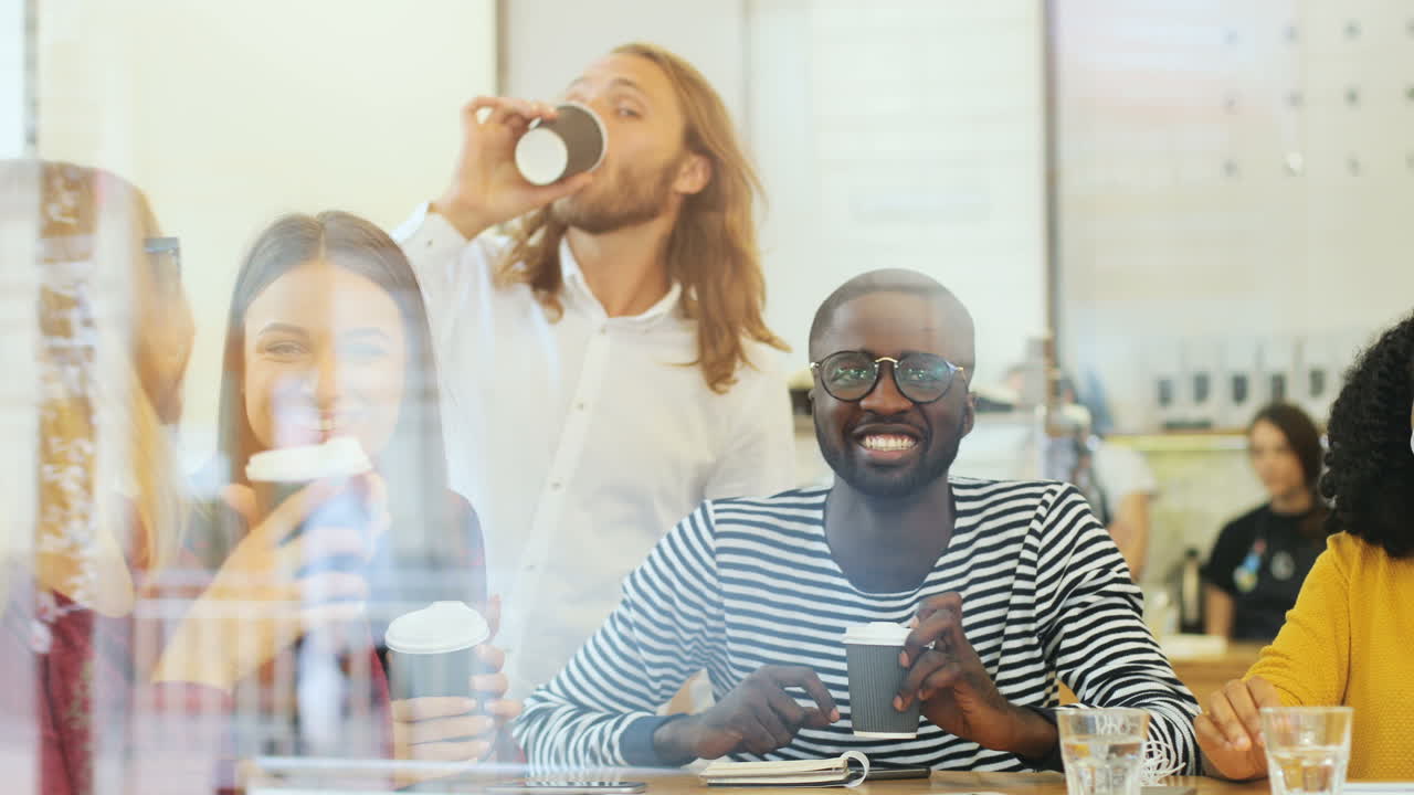 Camera focuses on a multiethnic group of friends through the window toasting with coffee cups sitting at a table in a cafe