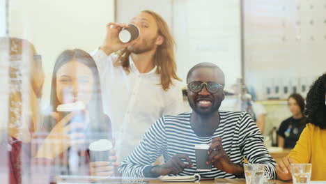 camera focuses on a multiethnic group of friends through the window toasting with coffee cups sitting at a table in a cafe