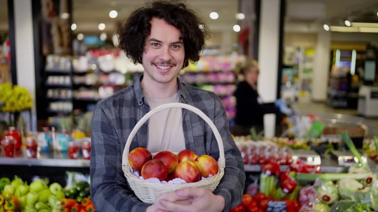 Portrait of a happy brunette guy with a mustache holding a basket of red peaches in his hands and posing in a grocery store
