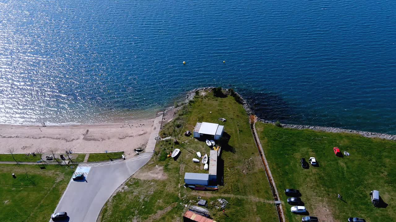 gente en el lago con jardín para caminar, playa y solarium con césped, barcos y coches aparcados, día soleado