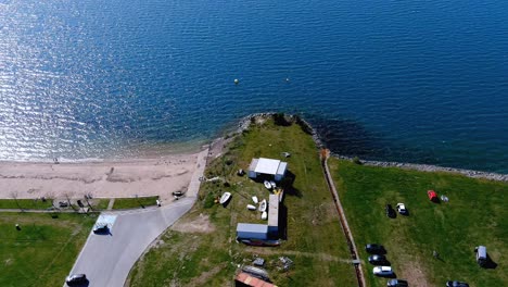 Menschen-Am-See-Mit-Garten-Zum-Spazieren,-Strand-Und-Liegewiese-Mit-Liegewiese,-Booten-Und-Geparkten-Autos,-Sonniger-Tag