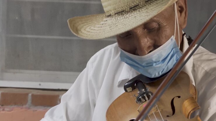 Primo piano di un anziano musicista ispanico con cappello e mascherina che suona il violino per strada.