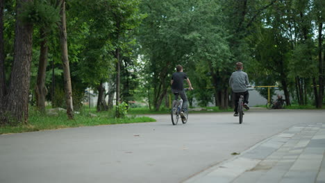 back view of two friends riding their bicycles along a park path, with lush green trees surrounding them and a blurred view of other people standing with bicycles ahead