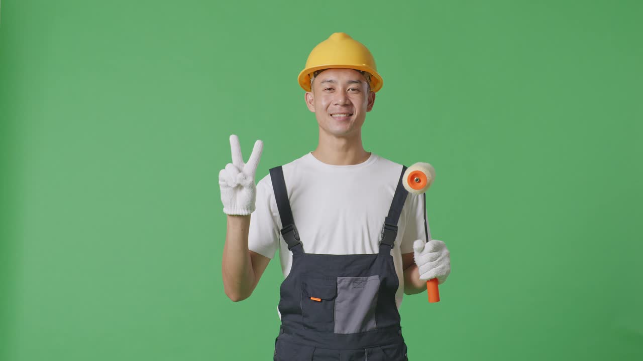 Asian Man Painter Wearing Safety Helmet Smiling And Showing Peace Gesture To Camera While Standing In The Green Screen Background Studio
