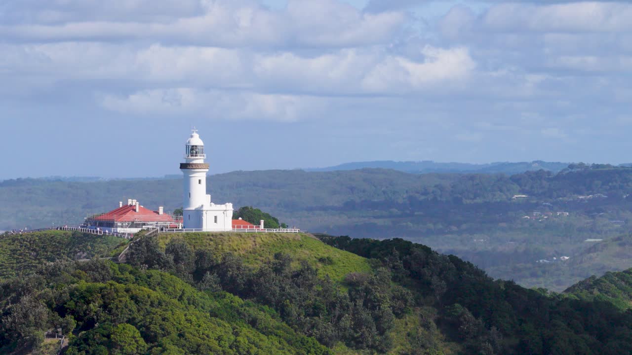 Drone captures Byron Bay lighthouse atop lush hills under a bright sky, offering a serene coastal panorama
