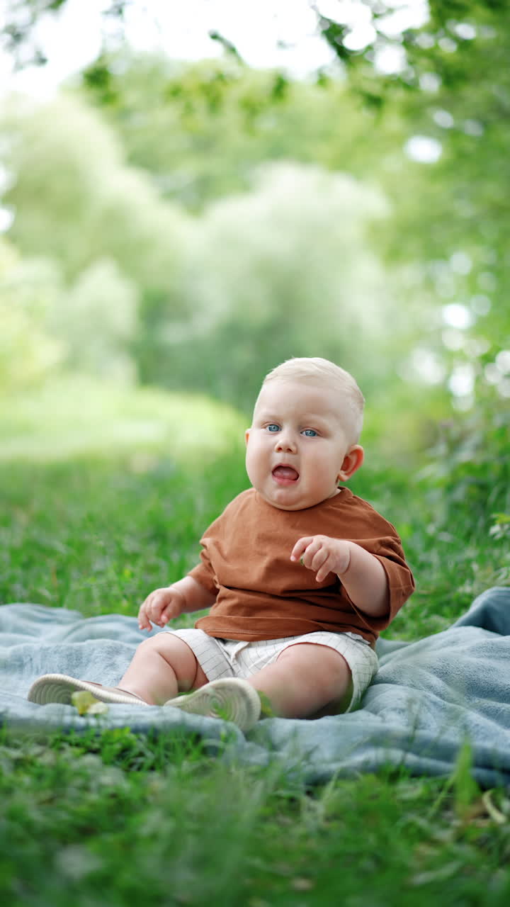 Calm cute Caucasian baby in brown t-shirt sits on the plaid. Cute child spending time outdoors in summer. Vertical video.