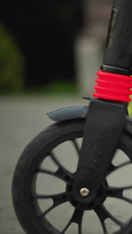 vista cercana de la pierna de una mujer montando un scooter negro, sus piernas visibles mientras se mueve hacia adelante, un niño pequeño sigue detrás en un scooter verde
