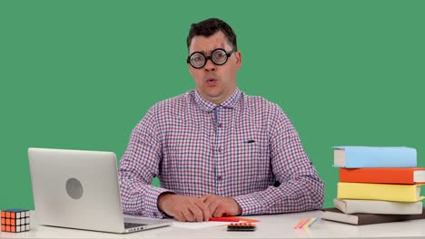 un hombre se sienta en un escritorio frente a una computadora portátil y libros, piensa en algo, asente con la cabeza negativamente, no hay ideas. un hombre con gafas en el estudio en una pantalla verde. cámara lenta. cerrar
