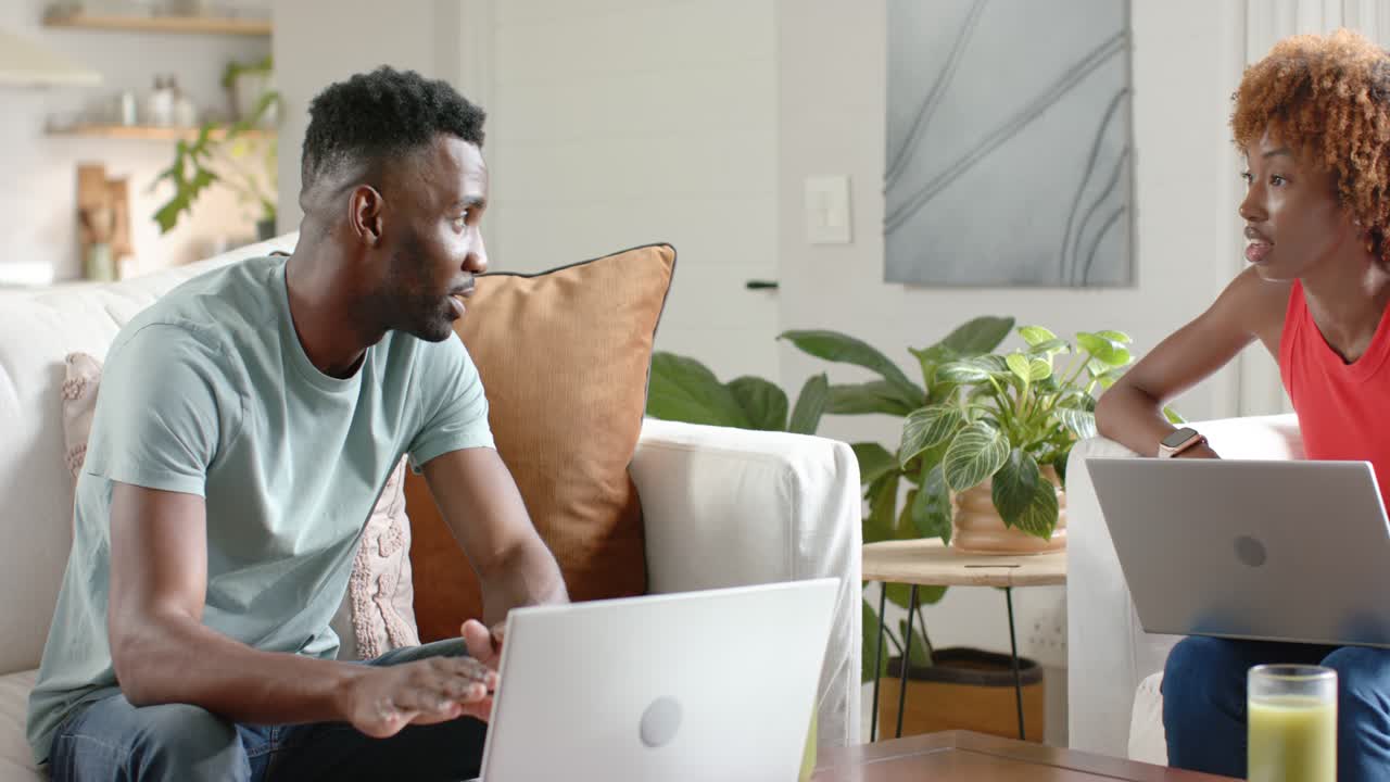 Diverse couple with laptops sitting at home, man typing and gesturing, woman listening and working