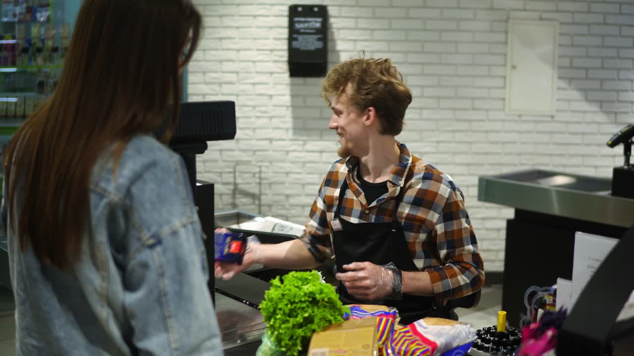 apuesto hombre joven barbudo escaneando comestibles, productos de caja en el mostrador. positivo, trabajador sonriente sirve a la mujer de pelo largo cliente irreconocible