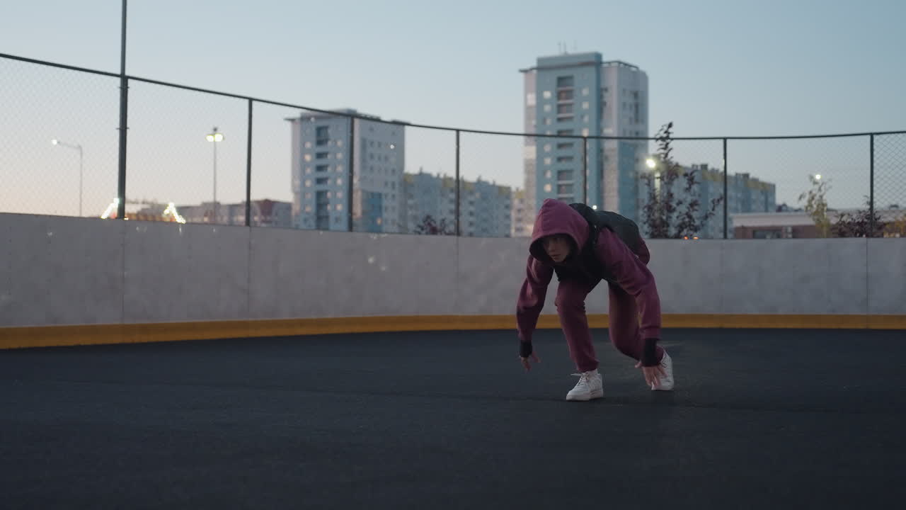 Female runner on white court line getting set to take off at dusk on black asphalt sports court near white barrier topped with chain link fence in urban setting with tall building backdrop
