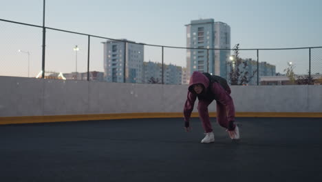 female runner on white court line getting set to take off at dusk on black asphalt sports court near white barrier topped with chain link fence in urban setting with tall building backdrop