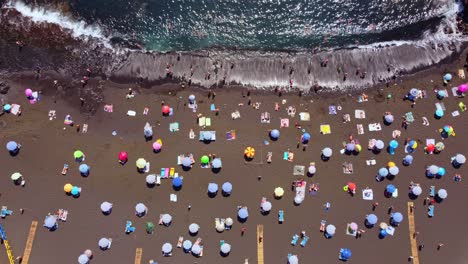 Top-down-aerial-view-of-tourists-sunbathing-on-the-black-volcanic-sand-beach-with-ocean-waves-touching-shoreline,-Play-de-la-arena,-Santiago-Del-Teide,-Tenerife-Island,-Spain