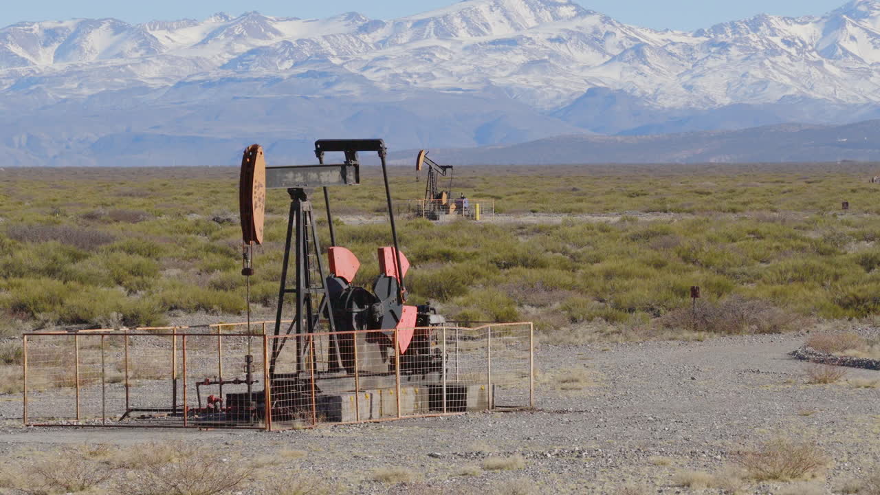 Oil pumpjacks operating in the arid steppe of Malargüe, Argentina, with the snow-capped Andes Mountains in the background.