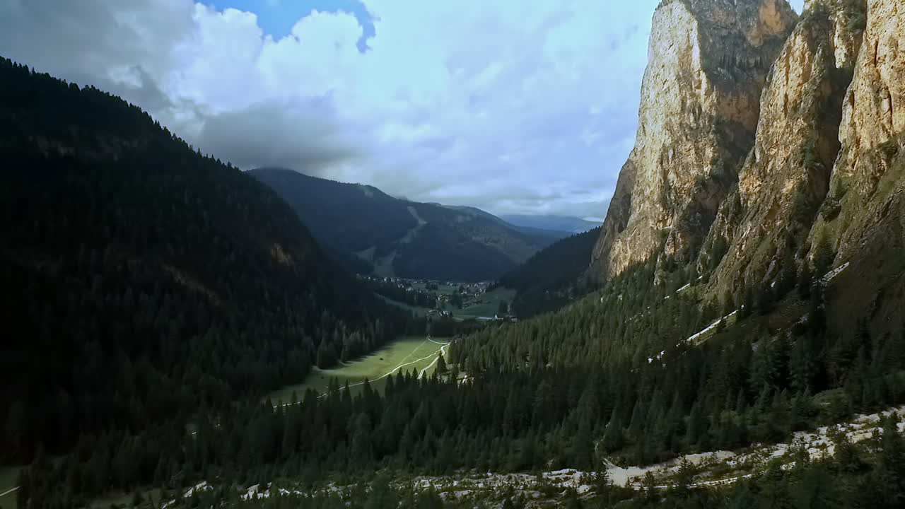 Sideways flight right to left across a beautiful alpine mountain valley with sheer rock on the right and wooded hills on the left