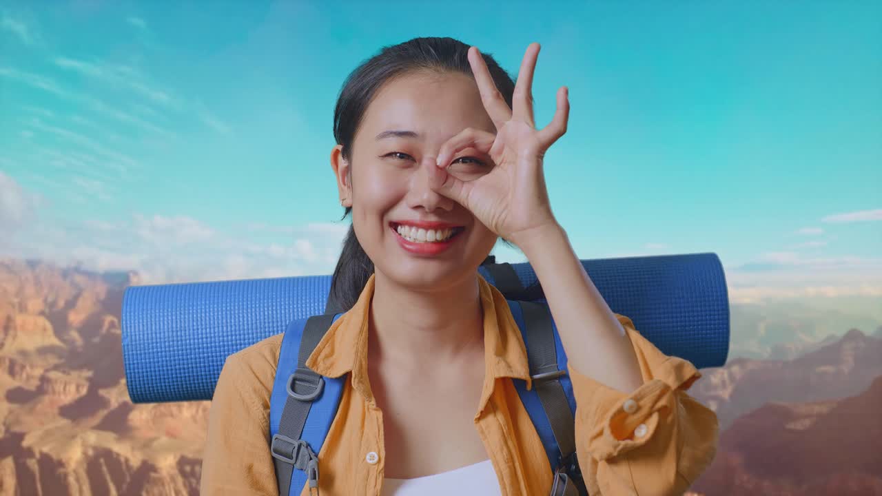 Close Up Of Asian Female Hiker With Mountaineering Backpack Smiling And Showing Ok Sign With Fingers Over Eyes While Traveling At The Top Of Mountain