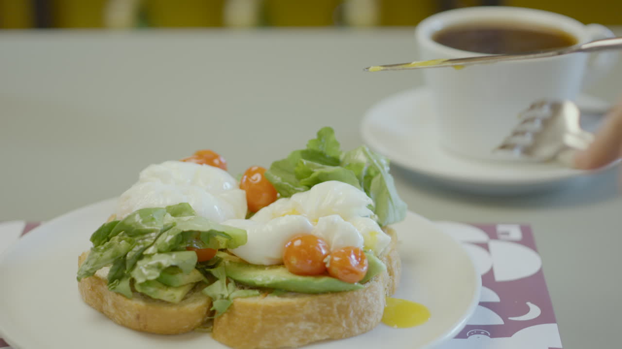 Close-up of a plate with rustic bread toast topped with avocado, lettuce, cherry tomatoes and poached eggs, next to a cup of hot coffee