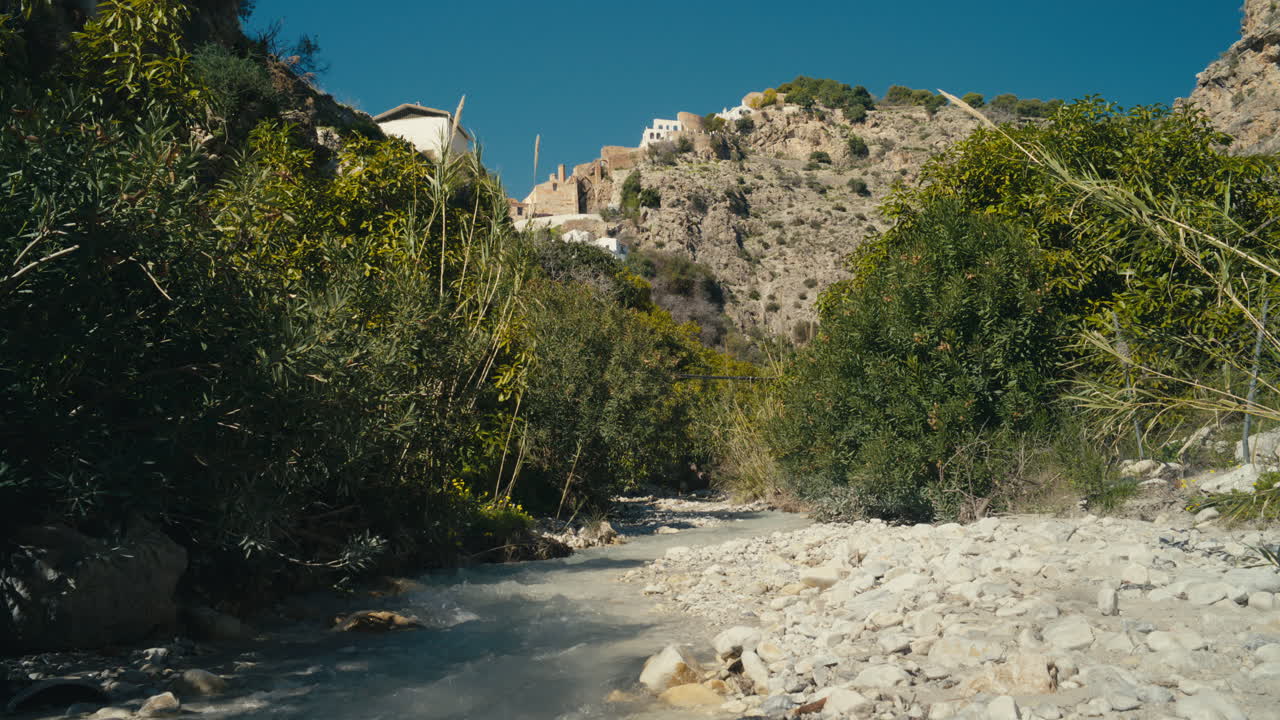 Chiaro ruscello di montagna che scorre attraverso un canyon roccioso circondato dalla vegetazione, con un villaggio collinare e antiche rovine in pietra sopra. Fusione scenografica di natura e cultura in un paesaggio mediterraneo.