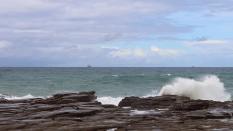 Olas-Rompiendo-En-Las-Rocas-Mientras-Un-Buque-Portacontenedores-Flota-En-El-Horizonte-Cerca-De-Sydney,-Australia.