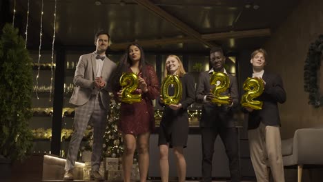 group of friends wearing elegant clothes holding balloons with the numbers 2022 while dancing and hugging each other in new year's party