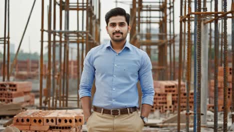 young indian engineer posing with hands in pockets at construction site, wearing shirt and trousers, smiling and looking at camera, with metal poles and bricks in background