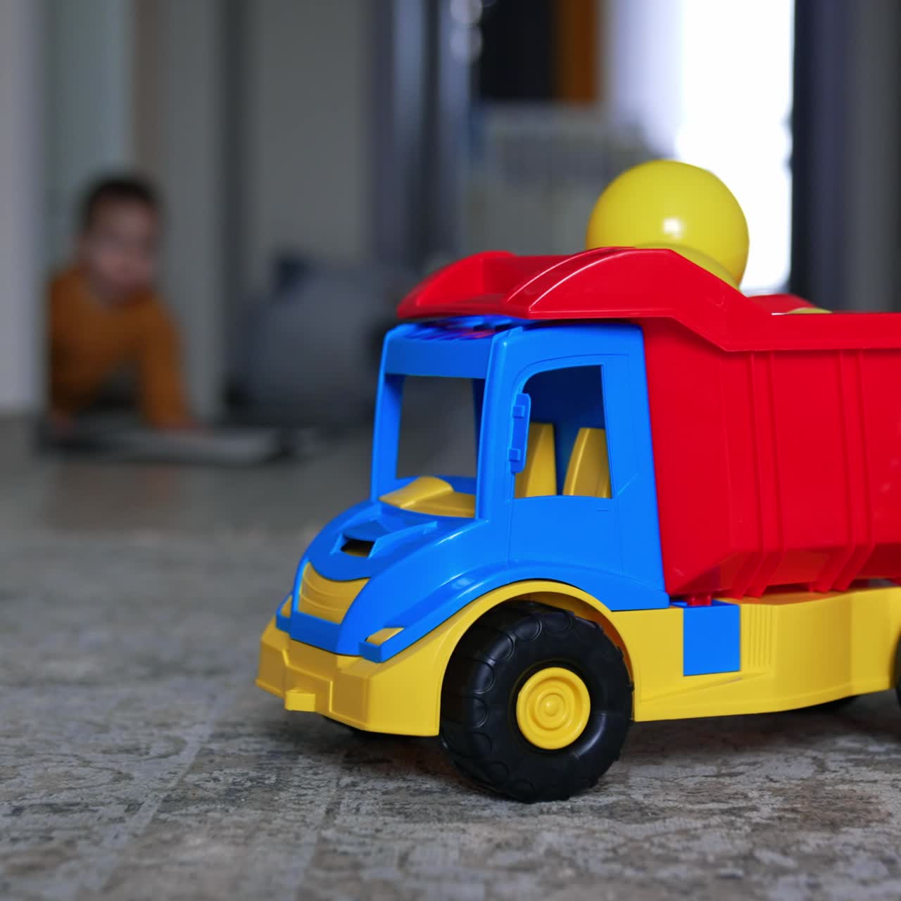 Colorful toy lorry on the floor of the room. Mom's hand puts the plastic balls on the car. Crawling child appears at backdrop in blur