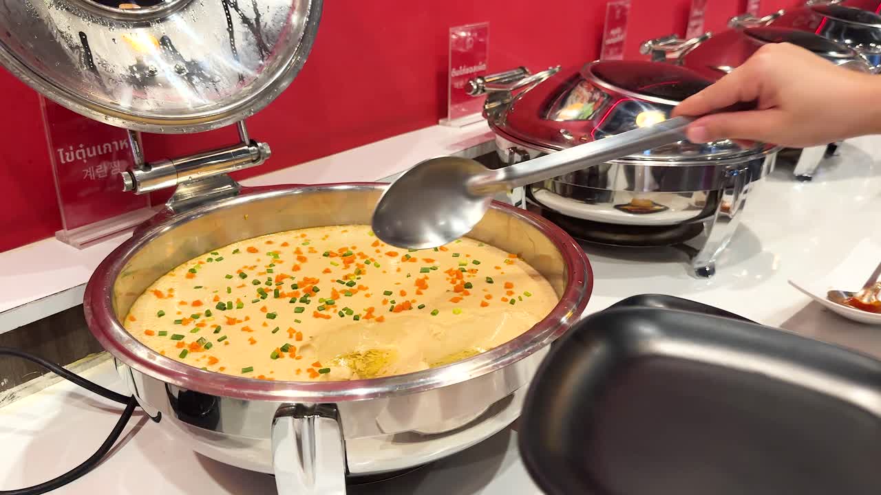 A person serves Korean steamed egg from a buffet station in a well-lit, vibrant setting