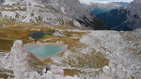 Toma-Aérea-De-Un-Dron-Sobre-Un-Modelo-Masculino-Parado-En-Rocas-Cerca-De-Tre-Cime-Di-Lavaredo-En-Los-Dolomitas,-Italia