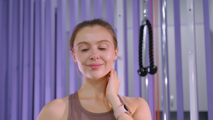 Donna con capelli castani raccolti che sorride calorosamente alla telecamera mentre si tocca la testa, in piedi con sicurezza davanti a uno sfondo viola.