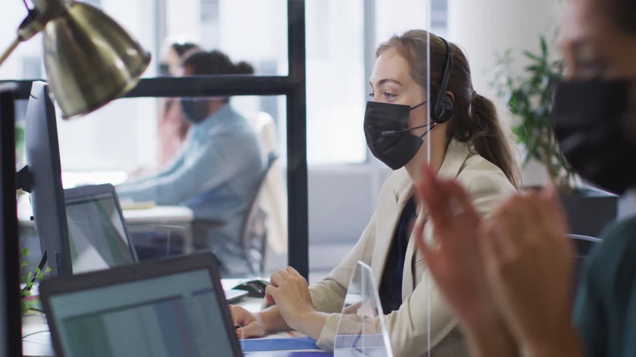 카우카시안 사업가 여성은 휴대폰 헤드 ⁇ 을 착용하여 재채기 방패와 함께 책상에서 얼굴 마스크를 착용하고 있습니다.