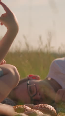 a boy wearing glasses and a white shirt lies on a scarf in a grassy field, gently touching a piece of bread. beside him, his younger brother, also in a white shirt, touches his nose in a thoughtful