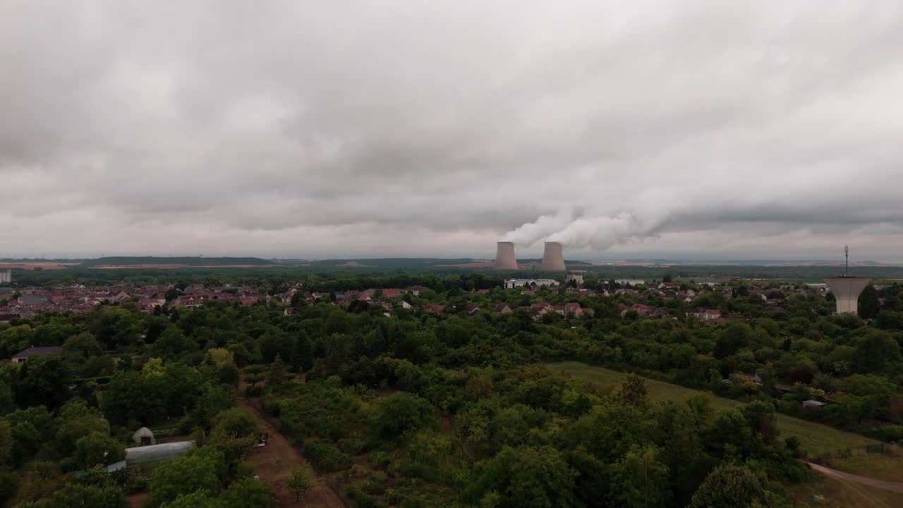 Aerial drone view of a French village with a nuclear power plant in the background. Cooling towers emit steam under a cloudy, overcast sky, showing a contrast between rural life and industrial energy