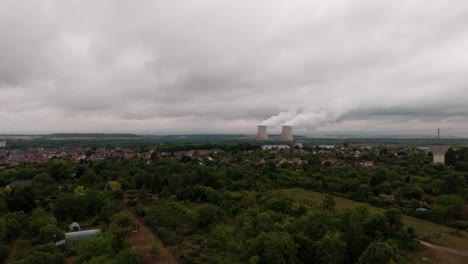 aerial drone view of a french village with a nuclear power plant in the background. cooling towers emit steam under a cloudy, overcast sky, showing a contrast between rural life and industrial energy