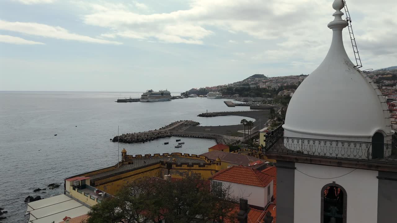Slowmotion aerial footage featuring Igreja do Socorro in Funchal, Madeira, with view over rooftops, harbor, cruise ships and Atlantic Ocean under cloudy skies