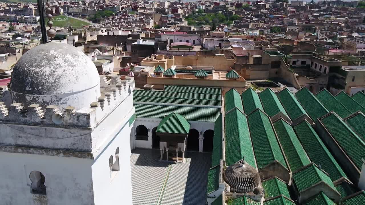 Exquisita toma de dron enfocada en el patio interior y el mihrab de Al-Qarawiyyin. Presenta el minarete blanco, techos verdes estampados y la disposición arquitectónica espiritual en la medina de Fez.