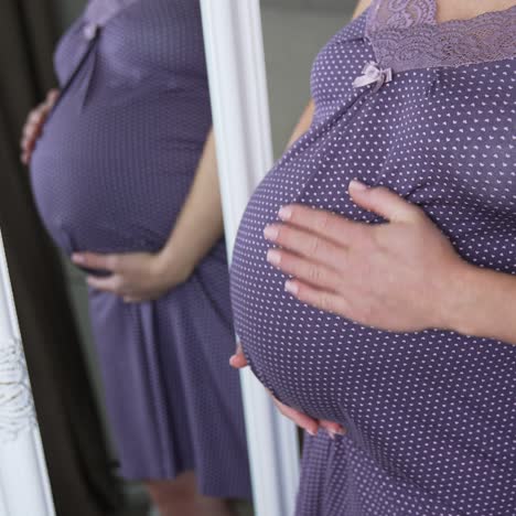 woman waiting for child stands in a purple nightshirt in front of the mirror. pregnant woman strokes her stomach with one hand reflecting in the mirror