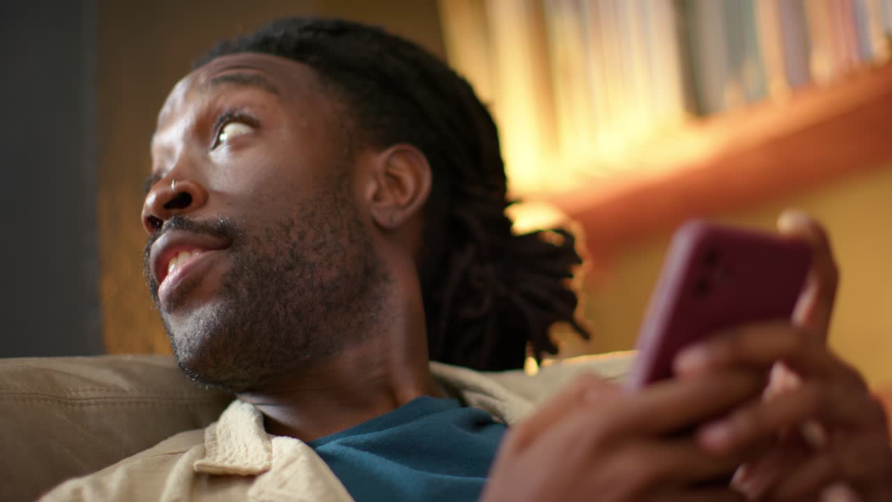 Uomo afroamericano che risponde a una notifica, toccando e scorrendo uno smartphone color malva sul divano.
