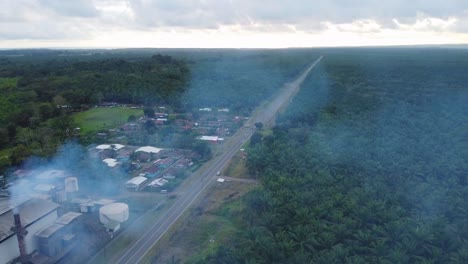 A-factory-near-a-palm-plantation,-aerial-drone-shot-of-the-building's-roof-with-a-steaming-chimney,-angled-forward-movement