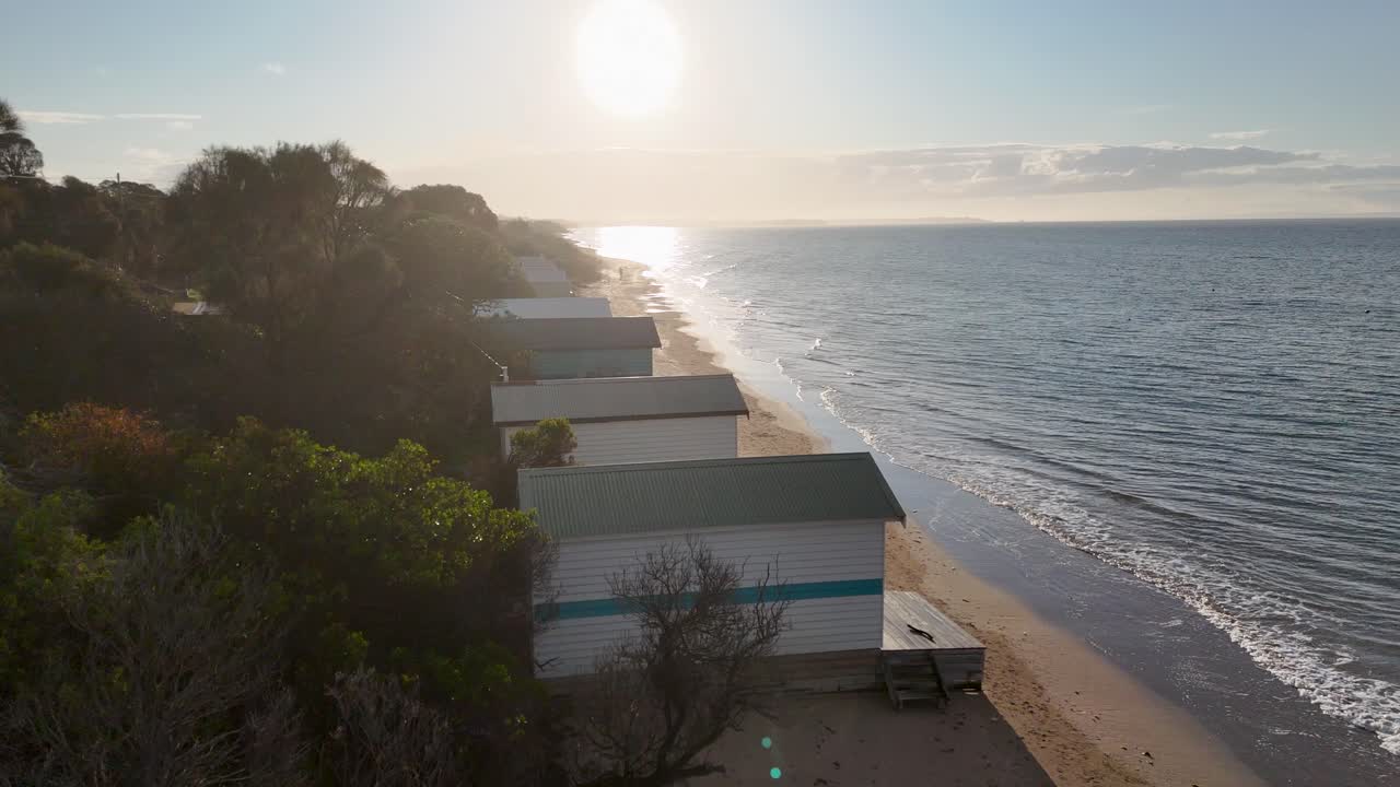 Drone glides above colorful beach huts at sunrise, capturing tranquil shoreline and gentle ocean waves