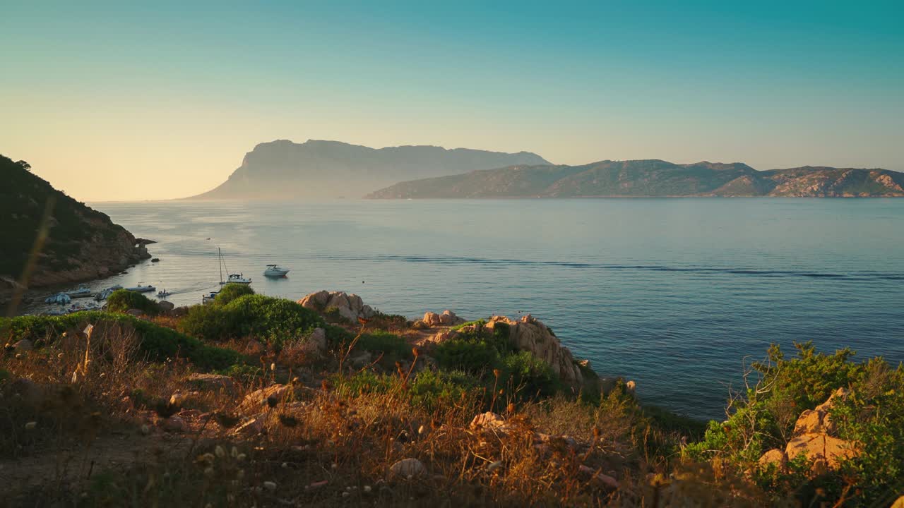 Kinemagrafi / saumaton videosilmukka purjeveneistä Italian turistilomasaarella Sardiniassa auringonlaskun aikaan, uimassa idyllisessä hiekkarannalla kirkkaan sinisen turkoosin ja tyynen veden äärellä lähellä Isola Tavolaraa ja Olbiaa. Drone-ilmakuva.