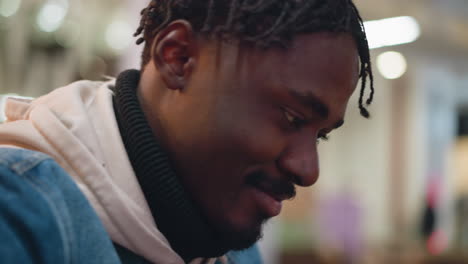 close-up profile of young black man indoors wearing denim jacket hoodie looking down smiling warmly with braided hair against blurred modern cafe background conveying relaxed thoughtful moment