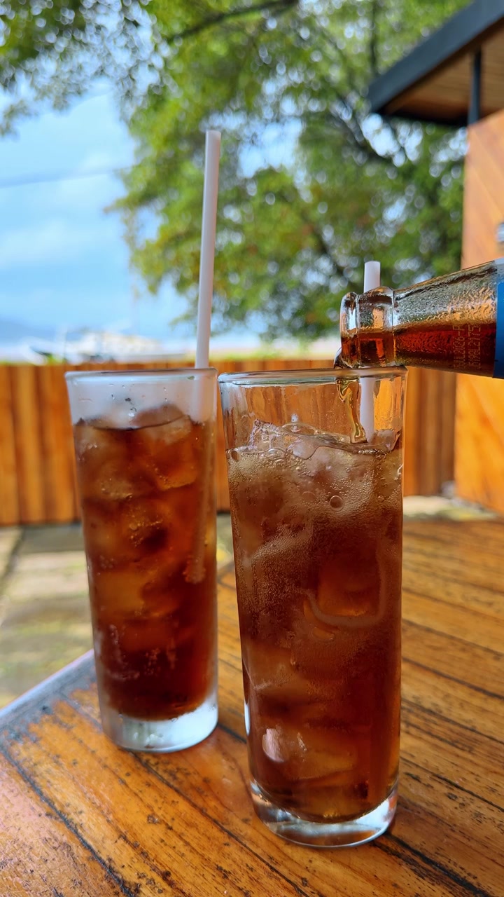 San José, Costa Rica, 1 de agosto de 2025: Dos vasos de refresco oscuro con hielo sobre madera. Vertiendo cola de una botella en vasos altos con cubitos de hielo sobre una mesa de madera en Costa Rica