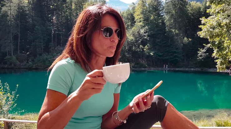 Donna anziana sportiva e attraente che si rilassa in riva a un lago immersa nella natura dopo aver fatto esercizio in estate e beve un caffè