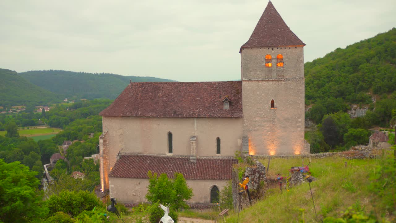 Wide shot of the church in Saint-Cirq Lapopie lit with warm lights during a cloudy afternoon in the French countryside