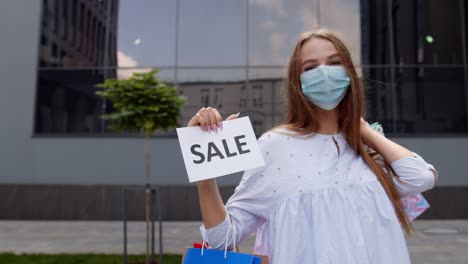 Girl-in-protective-mask-with-shopping-bags-showing-Sale-word-inscription-during-coronavirus-pandemic